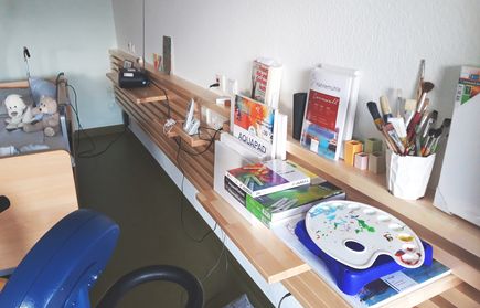 Wandregal mit Malmaterialien, Palette und Pinseln neben Bett mit Kuscheltieren