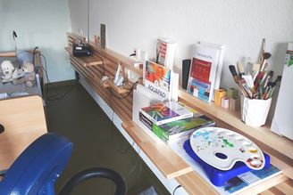 Wandregal mit Malmaterialien, Palette und Pinseln neben Bett mit Kuscheltieren