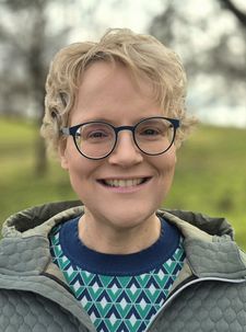 Lächelnde Person mit kurzen blonden Haaren und runder Brille