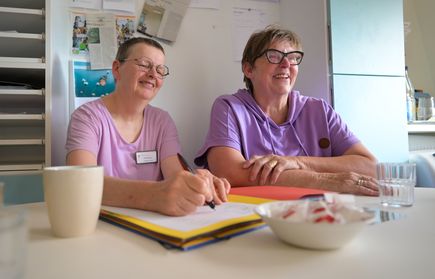 Zwei lächelnde Frauen in lila Kleidung am Tisch eine schreibt