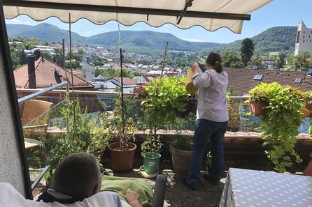 Person gießt Balkonpflanzen, Aussicht auf Ortschaft und bewaldete Hügel
