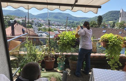 Person gießt Balkonpflanzen, Aussicht auf Ortschaft und bewaldete Hügel