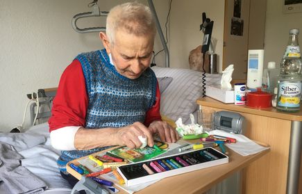 Älterer Mann sitzt am Bett und malt mit Buntstiften
