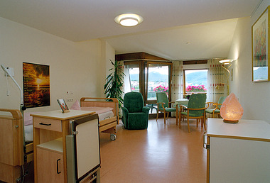 Helles Pflegezimmer mit zwei Betten und Sitzgruppe vor Fenstern