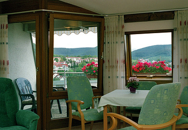 Wohnzimmer mit Tisch und Stühlen, Balkon mit Blumenkästen und Hügelblick